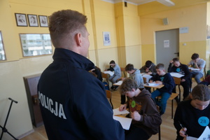 Na zdjęciu policjant, a w tle dzieci piszą test wiedzy.